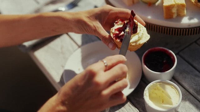 Hands spreading jam on a scone with cream and other treats