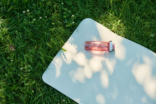 High angle view of fitness mat with reusable water bottle lying on green grass in park. Concept representing outdoor workout, wellness routine, healthy lifestyle marketing campaign