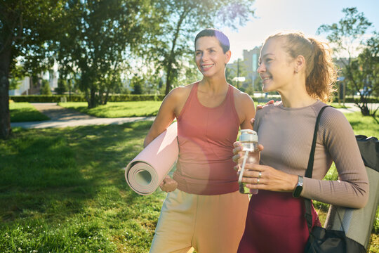 Middle aged Caucasian woman and young adult Caucasian woman holding yoga mats and water bottle walking outdoors in park. Suitable for fitness marketing and wellness campaigns