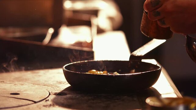 Chef stirs a small pan of food with two spoons in a dark kitchen