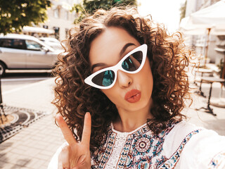 Beautiful smiling model with afro curls hairstyle dressed in summer hipster white dress.Sexy carefree posing in the street in sunglasses.Taking selfie self portrait photos on smartphone © halayalex