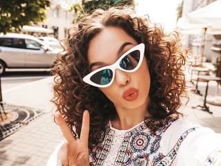 Beautiful smiling model with afro curls hairstyle dressed in summer hipster white dress.Sexy carefree posing in the street in sunglasses.Taking selfie self portrait photos on smartphone © halayalex