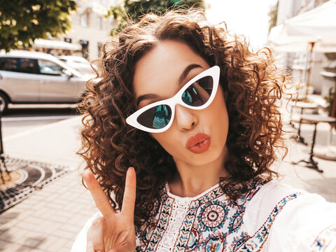 Beautiful smiling model with afro curls hairstyle dressed in summer hipster white dress.Sexy carefree posing in the street in sunglasses.Taking selfie self portrait photos on smartphone