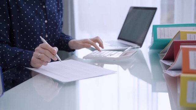A female accountant is using a laptop computer and calculator to calculate taxes at a glass desk in the office surrounded by vibrantly colored bookkeeping binders. Woman engaged in business audit and