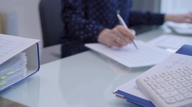 Calculator and binders of financial papers are waiting to be processed at the bookkeeper's workplace. A female accountant is using a laptop computer in the background. Woman engaged in business audit