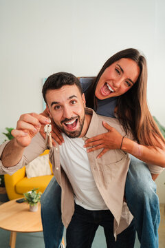 Two joyful lovers share a light moment as one shows a gleaming key toward the lens while the other hugs from behind, capturing energy and affection in a candid indoor scene. Close view shows warmth