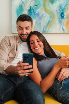 A smiling couple shares a quiet moment while viewing a smartphone screen. They lean close on a bright sofa, radiating warmth and connection as they explore content together in a cozy living room. now.