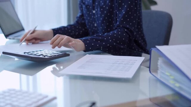 A female accountant is using a laptop computer and calculator to calculate taxes at a glass desk in the office. Woman engaged in business audit and finance