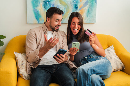 Two people sit close on a yellow sofa, smiling at their screens, sharing a card, and exploring online deals with excited teamwork and lighthearted digital browsing energy.