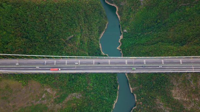 Aerial pull back view overlooking a vast highway suspension bridge spanning across a deep mountain canyon and river on an overcast day.