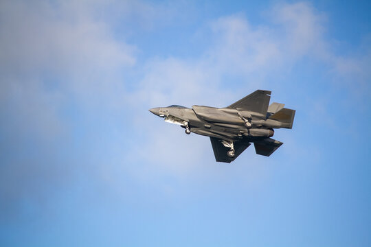F-35A Lightning II fighter aircraft flying across a blue sky with wheels down, photographed in side profile showing its distinctive stealth aircraft design.