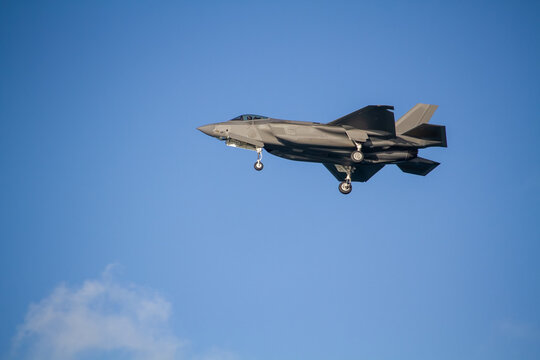 F-35A Lightning II fighter aircraft flying across a blue sky with wheels down, photographed in side profile showing its distinctive stealth aircraft design.