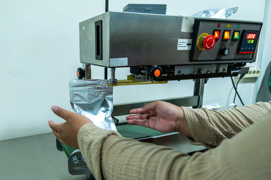 Worker hands operating industrial packaging machine sealing aluminum foil pouch in clean production environment.
