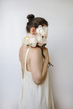 Brunette Woman In Beige Linen Dress Holding Light Pink Peonies Near Face