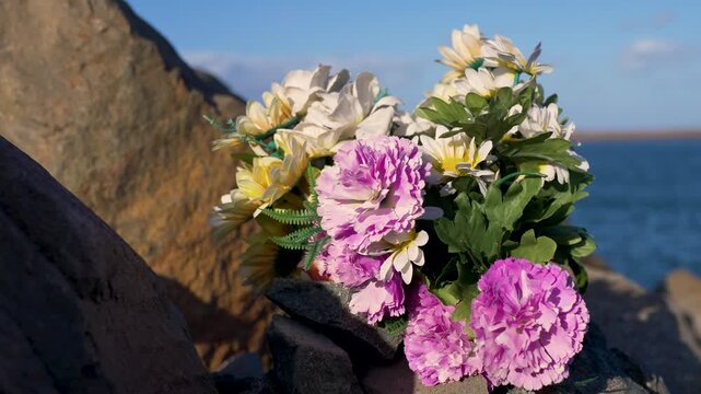 Slow motion close up of a colourful bouquet of artificial mixed commemorative flowers including pink carnations nestled among the rocks at the break wall waterfront pier in Newcastle city Australia