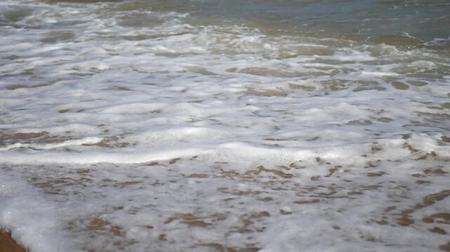 Close-up of sea waves washing over sandy beach with white foam moving across shallow water. Gentle ocean surf rolling onto the shoreline, natural seaside texture and relaxing coastal scene in daylight