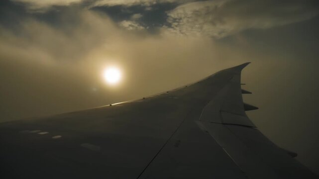 Airplane wing shaking during turbulence flying through thick clouds against bright sun. Passenger aircraft window view of bumpy flight in misty sky, strong wind, storm weather condition.