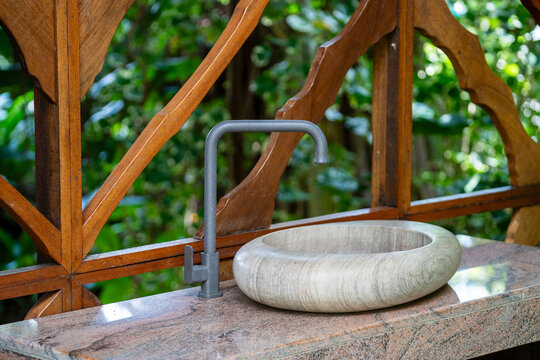 Modern stone washbasin and faucet in wooden outdoor interior. Stylish round stone vessel sink and gray matte faucet on granite countertop against wooden partition background in tropical garden