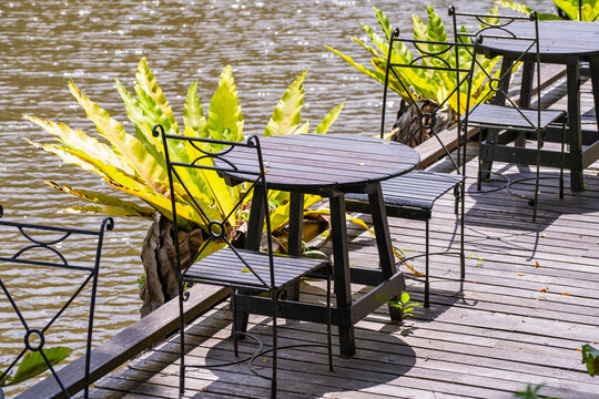 Outdoor cafe tables on wooden terrace by the water, close up. Wooden round tables and black wrought iron chairs on riverside deck terrace surrounded by lush green tropical plants in bright sunlight