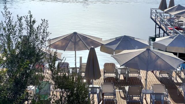 A sunny beach resort pier in Bitez, Turkey, with rows of white sun umbrellas and lounge chairs set up on a wooden deck over clear, calm sea waters.