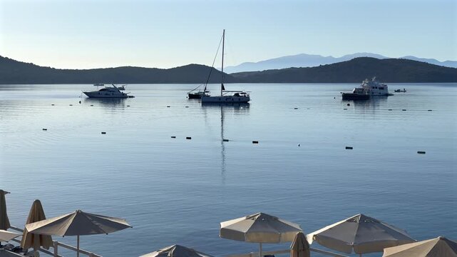 A tranquil view of Bitez Bay in Turkey, showing sailboats and yachts anchored on still, calm blue waters with sun umbrellas in the foreground and mountains.