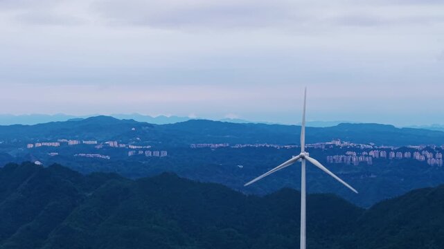 Aerial counterclockwise orbit of wind turbine on misty mountain ridge at cinematic twilight. Concept of renewable wind energy, eco sustainability, and carbon neutrality.