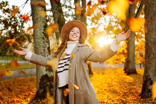 Woman wearing hat and trench coat is having fun in park covered with colorful leaves. Young woman enjoys autumn woods and tosses yellow leaves into air. Concept of leisure, weekends, and autumn.