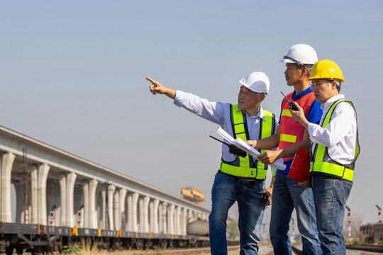 Team of engineers in hardhat pointing and discussing project at freight transport yard, industrial workers with blueprints and safety gear checking railway construction site and infrastructure