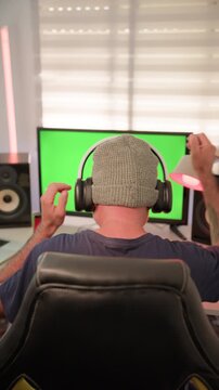 A man wearing a beanie sits in a gaming chair with his back to the camera, wearing headphones and dancing in front of a green screen monitor, Vertical