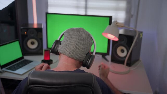 A man wearing a beanie touches a laptop, then puts on headphones with his back to the camera and dances in front of a green screen monitor and laptop