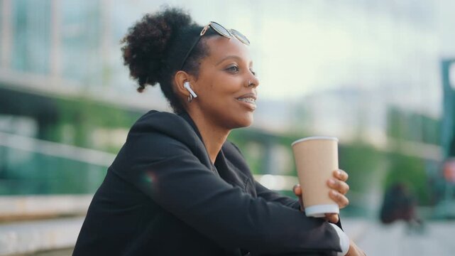 Joyful black woman with wireless headphones calling by mobile in city street. Portrait of modern townswoman with coffee cup in park, urban view and pretty lady, modern technology for communication