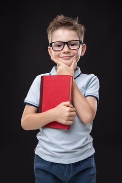 Smart schoolboy with eyeglasses holding red book thinking isolated on dark background