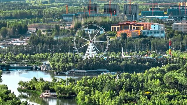 Aerial view of a Ferris wheel in a green city park by a lake with modern buildings