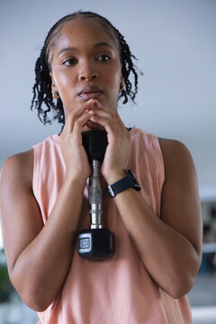 Adult African woman holding 2KG dumbbell at chest in gym, wearing pink tank, left-wrist watch
