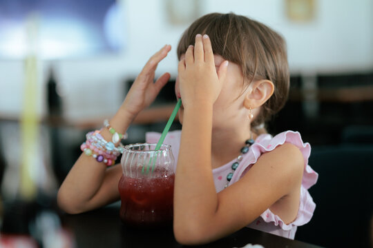 Playful Girl Hiding Her Eyes Eating Raspberry Lemonade Restaurant. Playful child covers her eyes, wearing colorful bracelets at a restaurant 