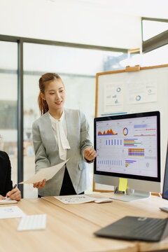 Asian businesswoman presenting financial data analytics on a computer monitor during a business meeting, explaining marketing strategy insights to colleagues in a modern office