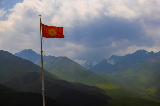 Vibrant red flag of Kyrgyzstan waves on a flagpole, set against rugged mountains. Dramatic clouds and sunlit hills enhance the natural beauty of the scene.