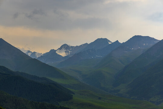 Wide scenic view of majestic mountain range under cloudy sky. Dark green forests transition to rolling hills, with a stream winding through the valley in Kyrgyzstan.