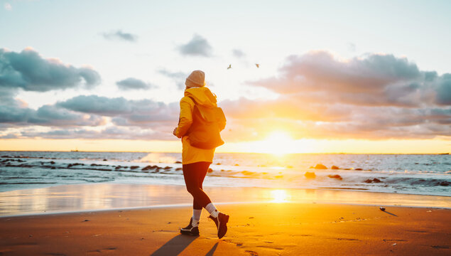 Traveler with backpack walks along sandy beach at sunset. Woman wearing bright raincoat and carrying backpack feels sense of freedom, enjoys seascape. Concept of adventure, freedom, and enjoyment.