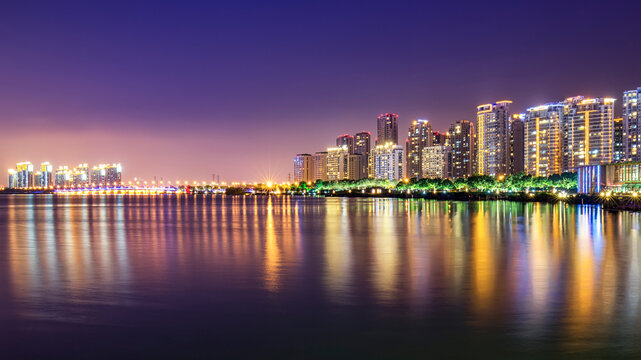 A calm body of water reflects vibrant city lights and high-rise buildings of an urban skyline during twilight.