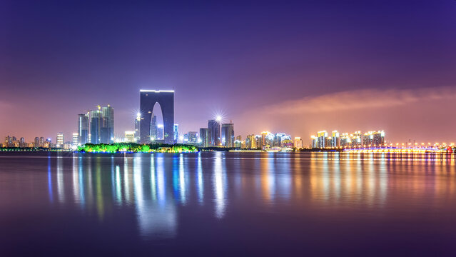 A modern urban night skyline featuring a distinctive arch-shaped skyscraper, with building lights reflecting on calm water under a dark purple sky.