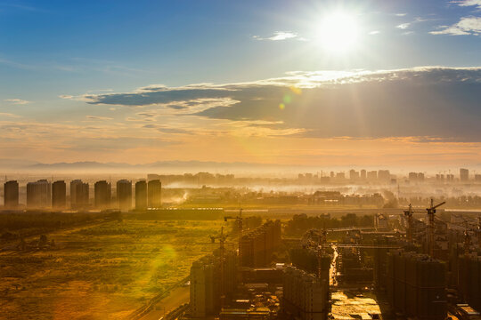 A misty urban landscape at sunrise, showing ongoing construction and distant city buildings under a bright sky.