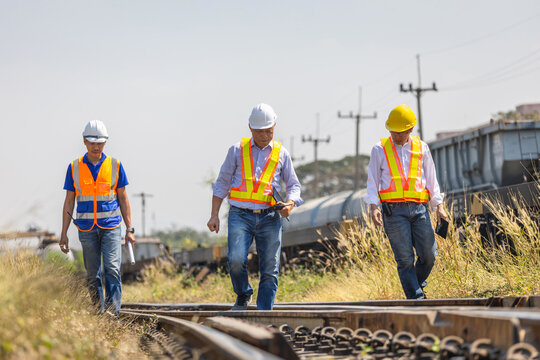 Team of engineers in safety vests and hard hats inspecting railway construction and transport infrastructure, industrial workers collaborating and walking together outdoors at freight yard facility