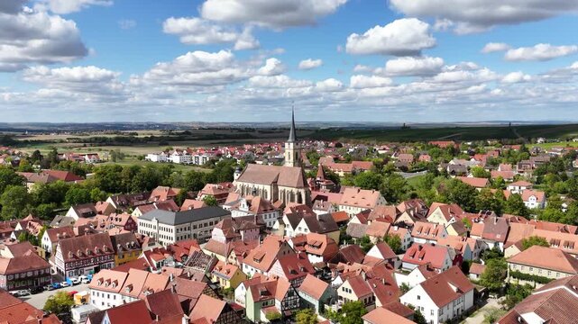 Malerische Panorama-Aussicht auf die historische Altstadt von Iphofen mit dem markanten Stadtturm, umgeben von gr&uuml;nen B&auml;umen unter einem blau-wei&szlig;en Sommerhimmel.