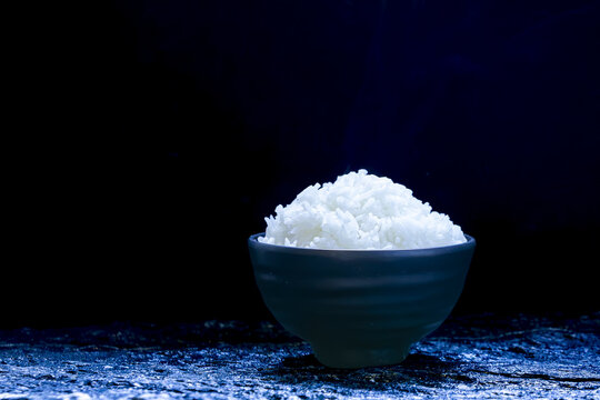 Steamed white rice in dark ceramic bowl, placed on textured surface with dramatic lighting, minimalistic food concept, Asian cuisine, healthy eating, simplicity, calm mood