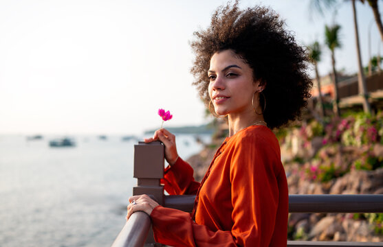 Portrait of a beautiful woman with curly hair holding a delicate flower by the sea at sunset
