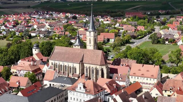 Malerische Panorama-Aussicht auf die historische Altstadt von Iphofen mit dem markanten Stadtturm, umgeben von gr&uuml;nen B&auml;umen unter einem blau-wei&szlig;en Sommerhimmel.