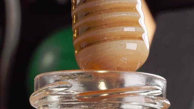 Honey dripping from wooden dipper into open glass jar, showing glossy golden liquid, viscous texture and slow flowing stream against dark background