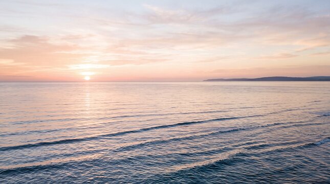 Beautiful pink and golden sunset over calm ocean waves with soft clouds and distant coastline mountains during peaceful evening reflection