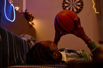 Young African American man lying down while holding basketball indoors. Concept image useful for...